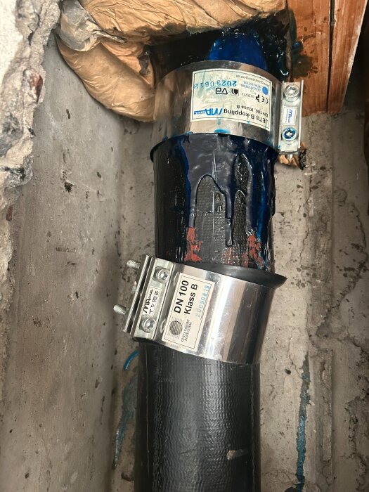 Svart rör med metallklämmor och packning mot en vägg, märken och etiketter synliga. Montaget sker i ett hål i en betongvägg.