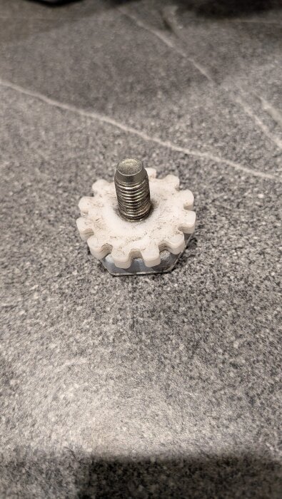 Löst maskinfot i plast och metall med gängad skruv, lagt på en grå stenyta. Skruvgängorna är synliga och plasten är delvis smutsig.