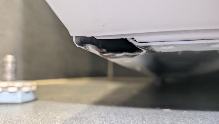 Skadat hörn av tvättmaskin med bortrostat eller bortbrutet metallparti där ett ben ska monteras.