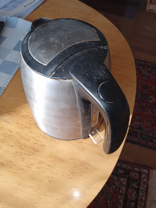 En använd vattenkokare med silverfärgad kropp och svart handtag på ett träbord.