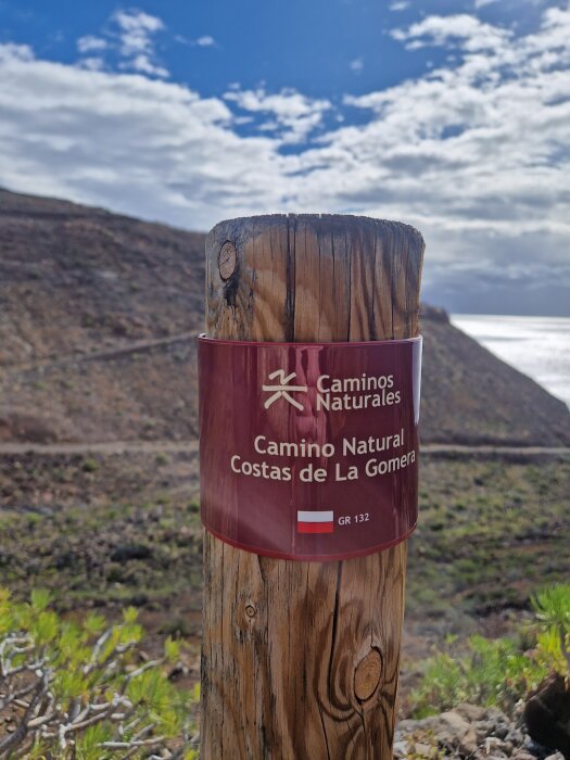 Trästolpe med röd skylt visar "Camino Natural Costas de La Gomera" mot bergig bakgrund och blå himmel.