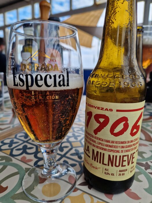 Glas och flaska med mörk öl från 1906 Cerveceros, 6,5% alkohol, på ett färgglatt bord med kafémiljö.