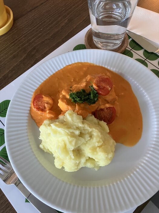 Tallrik med potatismos och kyckling i orange sås med tomat, bredvid ett glas vatten.