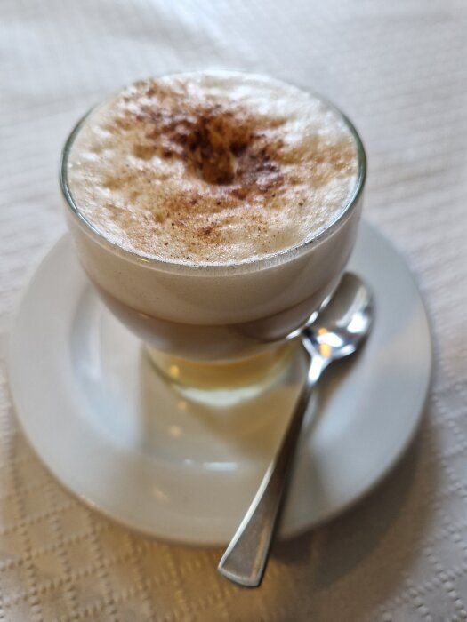 En glas med kaffedrycken Barraquito, toppad med skum av kondenserad mjölk och kanel, placerat på ett fat med sked.