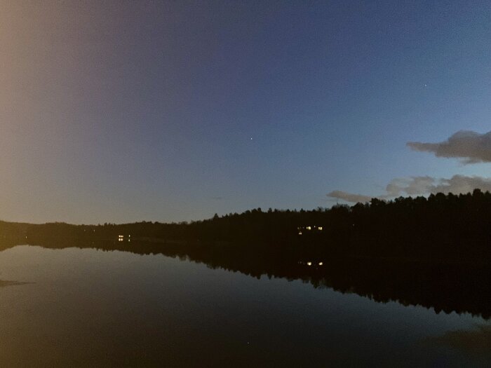 Kvällslandskap med stilla sjö som speglar skogen, himlen är skymningsblå och några ljuspunkter syns bland träden i fjärran.