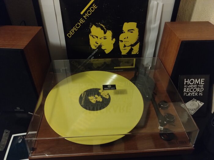 Gul vinylskiva på en skivspelare med Depeche Mode-omslag i bakgrunden och högtalare omkring.