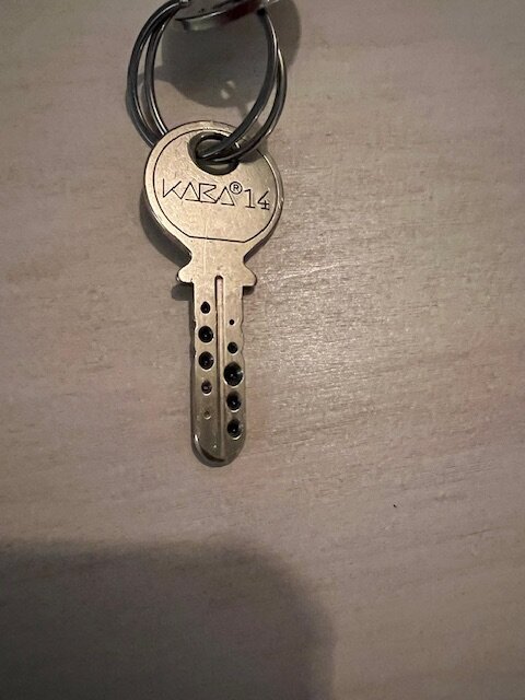Nyckel med texten "Kaba 14" och små gropar, hängande i en nyckelring, ligger på en bordsyta.