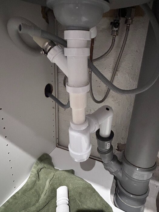 Vitt avloppssystem under diskbänk med rör och anslutningar, grön trasa och vit rördel synliga.