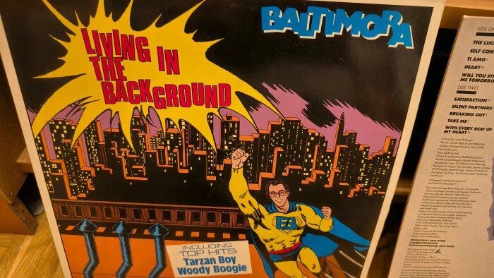Skivomslag från Baltimora med titeln "Living in the Background" och superhjälteillustration, innehållande låtar som "Tarzan Boy" och "Woody Boogie".