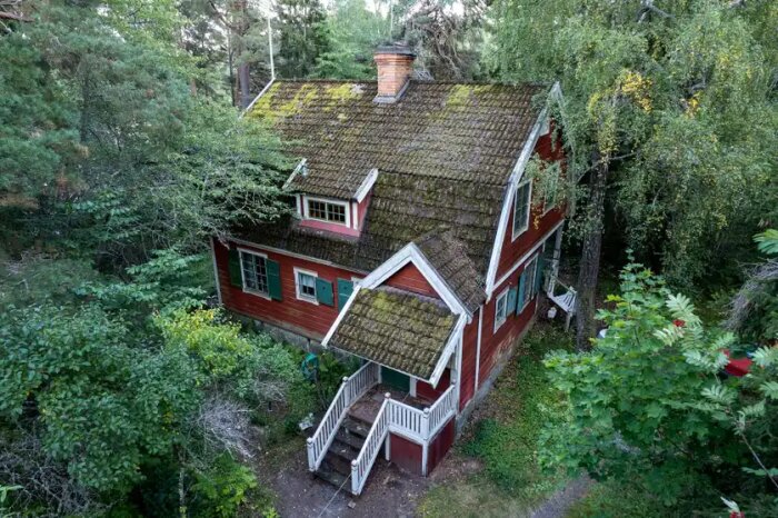 Gammalt rött trähus med mossa på taket omgivet av träd, potentiellt isolerat med direktverkande elvärme.