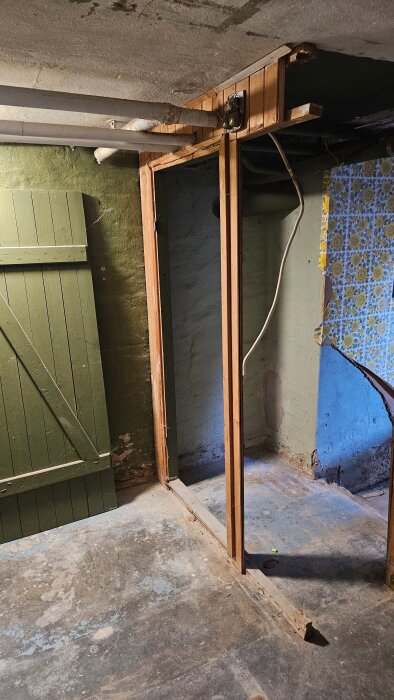 Träram och stålrör i källare, möjligen för skiljedörr, i hus från 1936. Grön dörr och blommig tapet syns på väggarna.