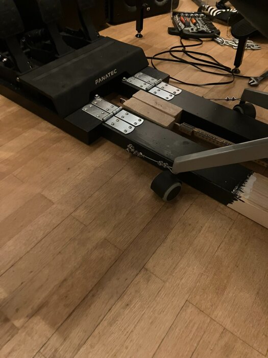 Pedalkonstruktion monterad på träreglar med gångjärn och stödhjul, på ljus trägolv. Verktyg ligger i bakgrunden.