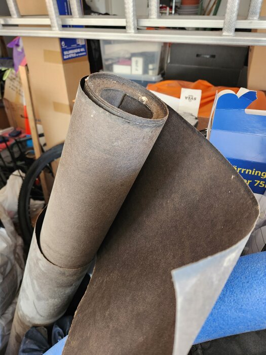 Rullad papp för takläggning ligger på en hylla i ett förråd fyllt med diverse byggmaterial och kartonger.
