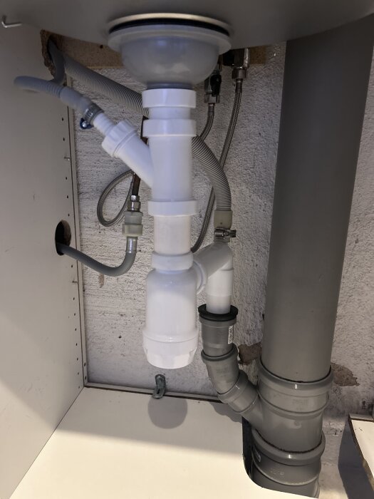 Under vasken installation av tvättmaskins- och kondenstumlarslangar. Grå slangar anslutna till avloppet, vit huvudledning.