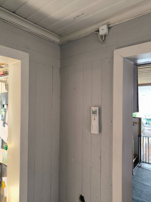 Insida av ett gammalt hus med plankväggar målade i ljusgrått och elkablar monterade längs taket. En termostat syns på väggen.