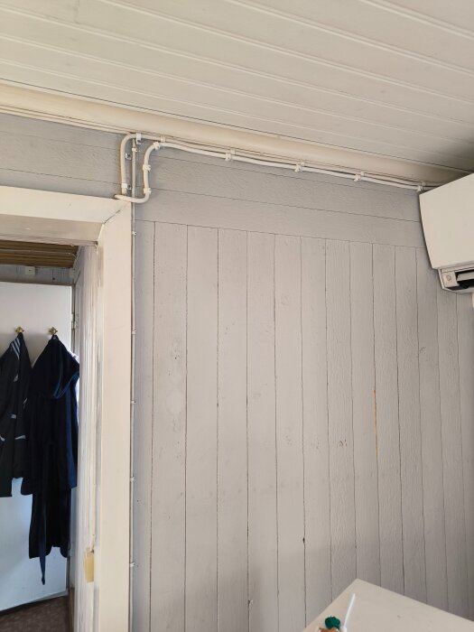 Plankvägg i ett äldre hus med synlig eldragning i plastslangar upptill, nära taket. En dörröppning och hängande kläder syns till vänster.