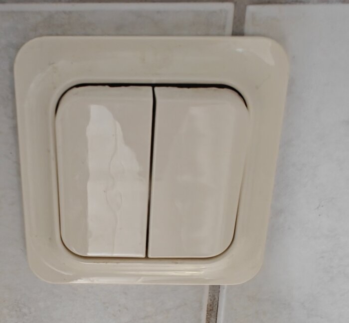 Dubbel ljusknapp i beige, monterad på en kaklad vägg i ett badrum.