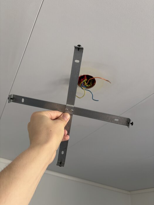 A hand holding a metal mounting bracket against a ceiling with exposed wiring from a hole, seeking advice on securing a lamp fixture.