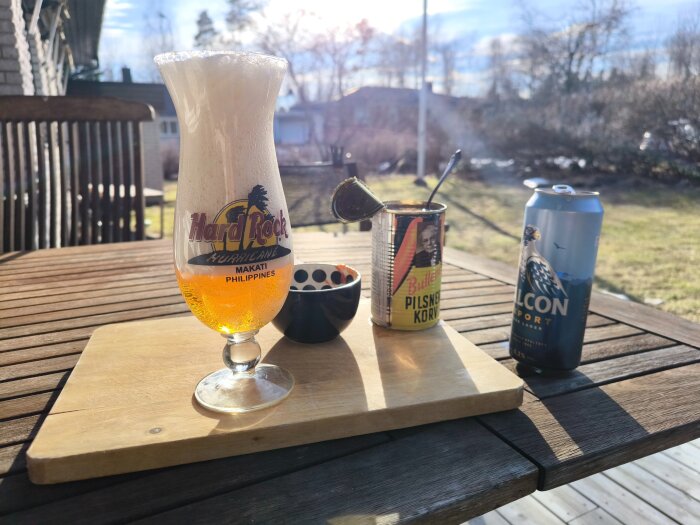 Öl i Hard Rock-glas, Bullens pilsnerkorvburk, Heinz hotsauce på skärbräda på en altan med gräsmatta i bakgrunden.