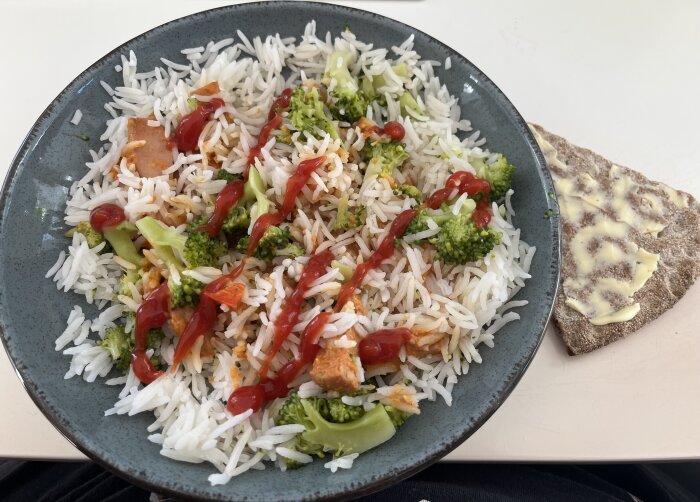 Tallrik med ris, broccoli och korvstroganoff toppat med ketchup, bredvid en smörad knäckebrödsskiva på vit bakgrund.
