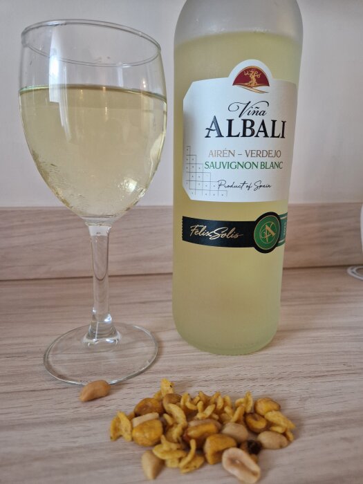 Flaska och glas med vitt vin bredvid en liten hög snacks på bord.