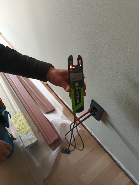 Hand håller en mätare vid ett vägguttag för att kontrollera jordning, medan en röd och svart sladd är ansluten till uttaget.