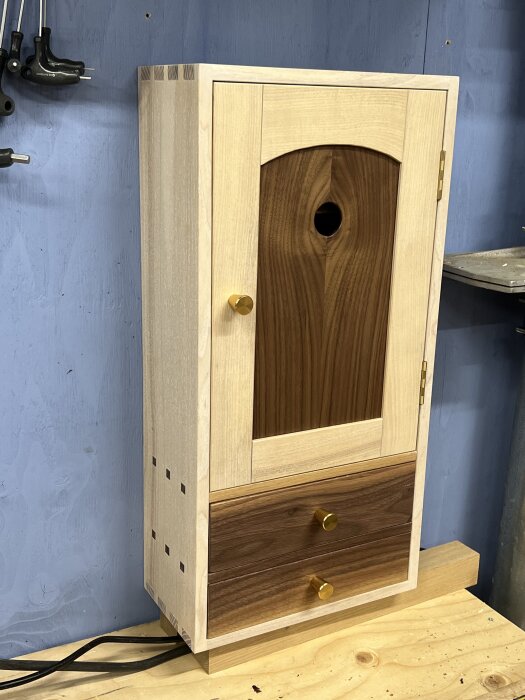 Träskåp med dörr, två lådor och gyllene knoppar, stående på en bänk i verkstadsmiljö, saknar upphängningsanordning.