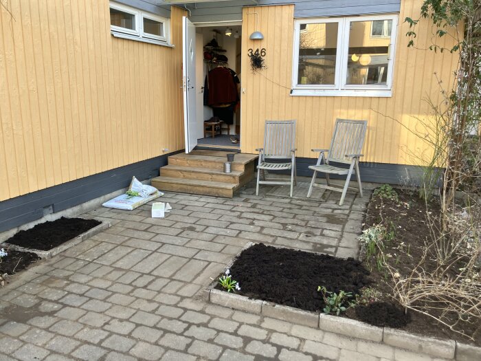 Gulmålad fasad med två stolar på uteplatsen, nyplanterade rabattområden och blomlökar som tittar upp ur jorden framför huset.
