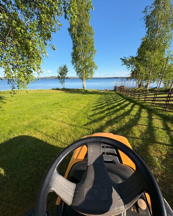 Åkgräsklippare på gräsmatta vid sjö med träd och trästaket i bakgrunden.