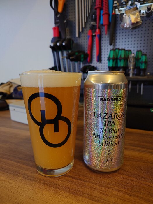 Ölglas med skummande dryck och en burk Lazarus IPA på ett bord, verktygstavla i bakgrunden.