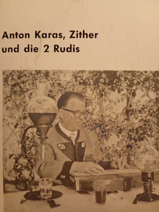 Anton Karas spelar ett stränginstrument, omgiven av gammaldags föremål, med titeln "Anton Karas, Zither und die 2 Rudis" i överkant.