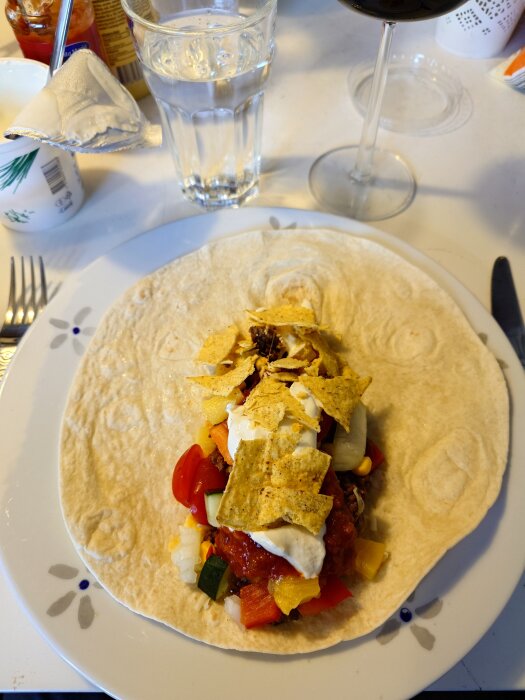 En tortilla fylld med tacos, grönsaker, sås och nachochips på en tallrik, omgiven av ett glas vatten och ett glas rödvin.