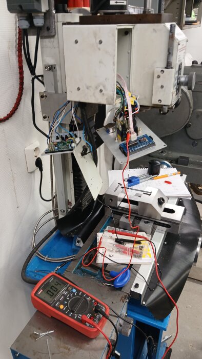Multimeter och elektronikkomponenter på ett arbetsbord intill en maskin i verkstadsutrustning, med kablar och anteckningsblock i bakgrunden.