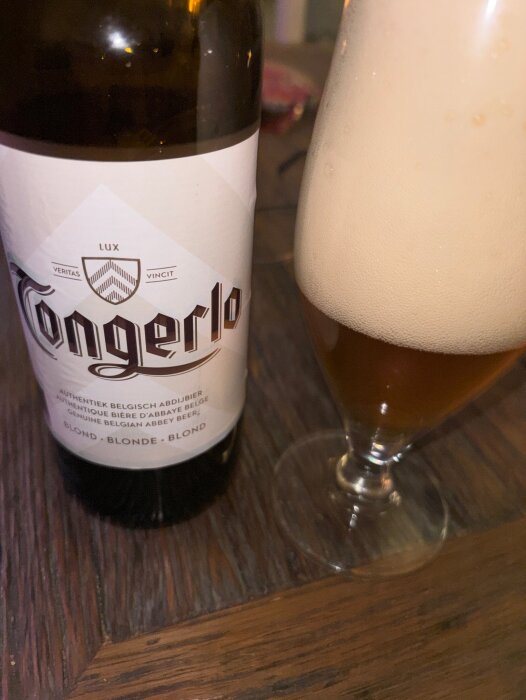 Belgisk abbeyöl av märket Tongerlo i en flaska och ett glas med skummande blont öl på ett träbord.