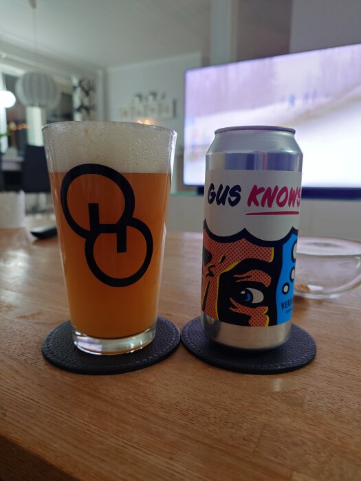 Ett ölglas med logotyp och en burk med etiketten "Gus Knows" på ett bord, med en bakgrund av ett vardagsrum och TV.