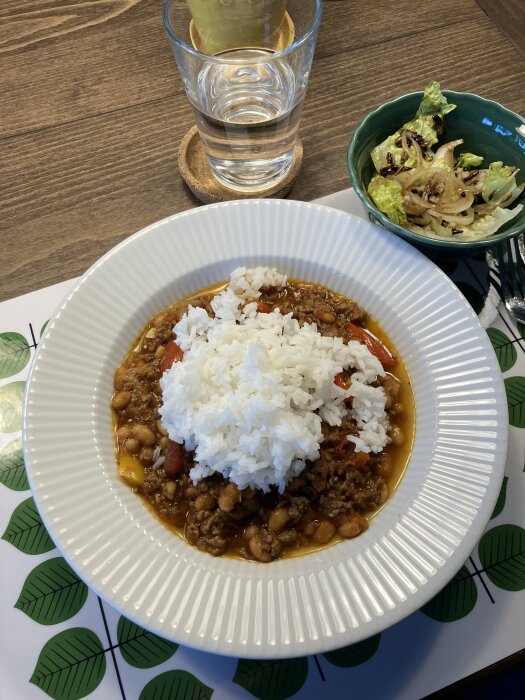 Brunt fat med chili con carne täckt av ris bredvid skål med isbergssallad och ett glas vatten på ett bord.
