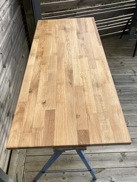 Lackat hyllplan av eklimfog som används som bänkskiva på köksö. Brädet har en slät, ljusbrun yta och är placerat på ett stafli utomhus.