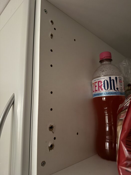 Skåpsinredning med flera borrhål, några skadade. En plastflaska står bredvid. Används för fråga om återmontering av skåplucka.