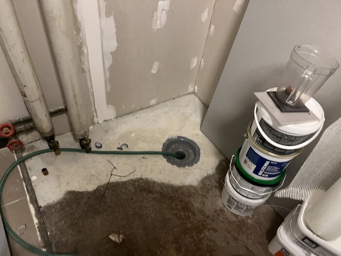 Pannrum under renovering med ny golvbrunn och betonggolv. Staplade burkar och verktyg syns, samt rör längs väggen.