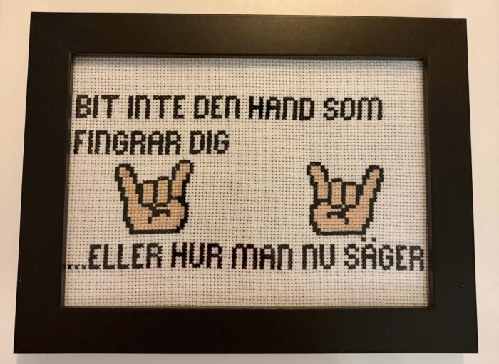 Inramat korsstygn med texten "BIT INTE DEN HAND SOM FINGRAR DIG...ELLER HUR MAN NU SÄGER" och två händer med rocktecken.