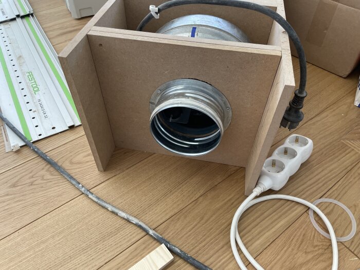 Luftfiltreringsenhet av trä, med rör och elkabel, placerad på trägolv och omgiven av verktygstillbehör.