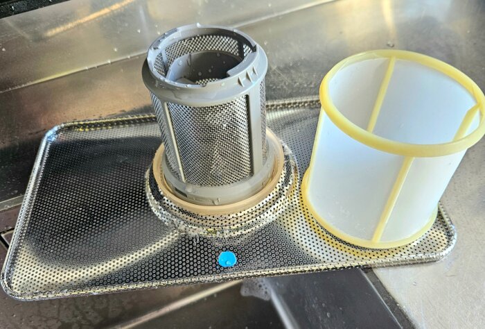 Två diskmaskinsfilter från Bosch, ett cylinderformat och ett cylindriskt vitt filter, placerade på en metallbricka med perforerade hål.