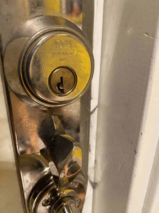 Närbild av en dörrlås av märket ASSA ABLOY på en vit dörr, visar låscylinder och vridhandtag i metall.