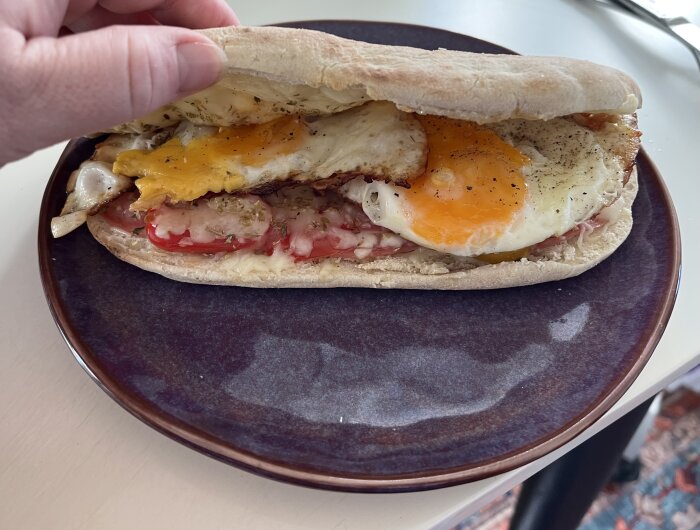 En hand håller en öppen smörgås på ett fat, med stekt ägg, smält ost, tomater och kryddor som fyllning.