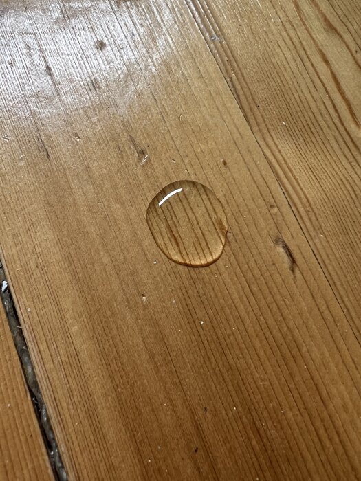 Vattendroppe på ett gammalt trägolv med märken och nyansskillnader, testas för att avgöra golvets behandling.