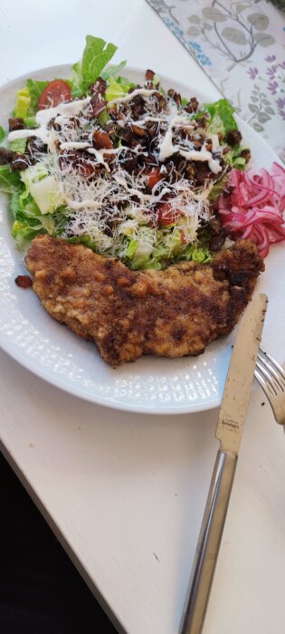 Ceasarsallad med schnitzel, toppad med parmesan, dressing och rödlök, serverad på en vit tallrik med bestick.