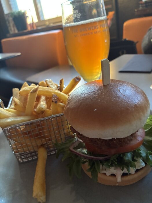 Crispy chicken-burgare med sallad, tomat och aoli på ett bord, bredvid pommes frites i en metallkorg, samt ett glas kall dryck i bakgrunden.