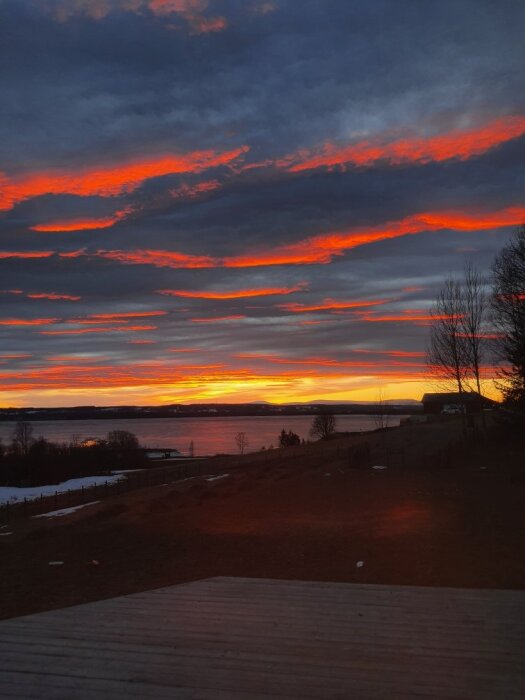 Solnedgång med intensivt röda och orange moln över en sjö, träd och ett hus i siluett i förgrunden.