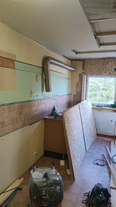 Kök under renovering med borttagna skåp, synliga ventilationsrör, byggmaterial och verktyg på golvet. Fönster med utsikt mot grönska.