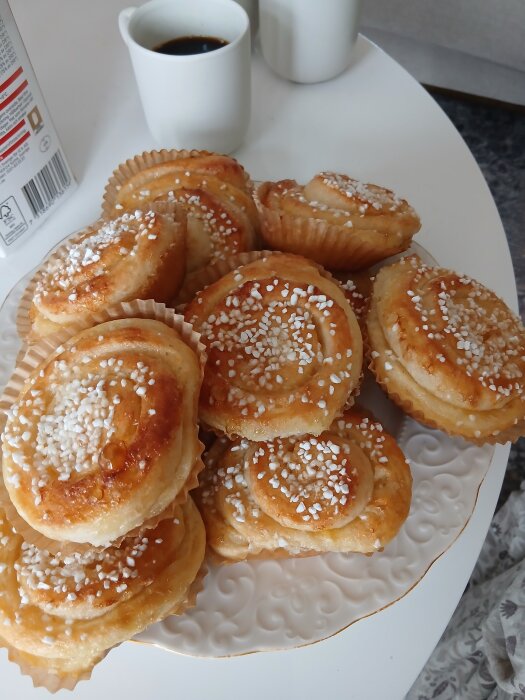 En tallrik med nybakade bullar toppade med pärlsocker, serverad bredvid en kopp kaffe på ett vitt bord.